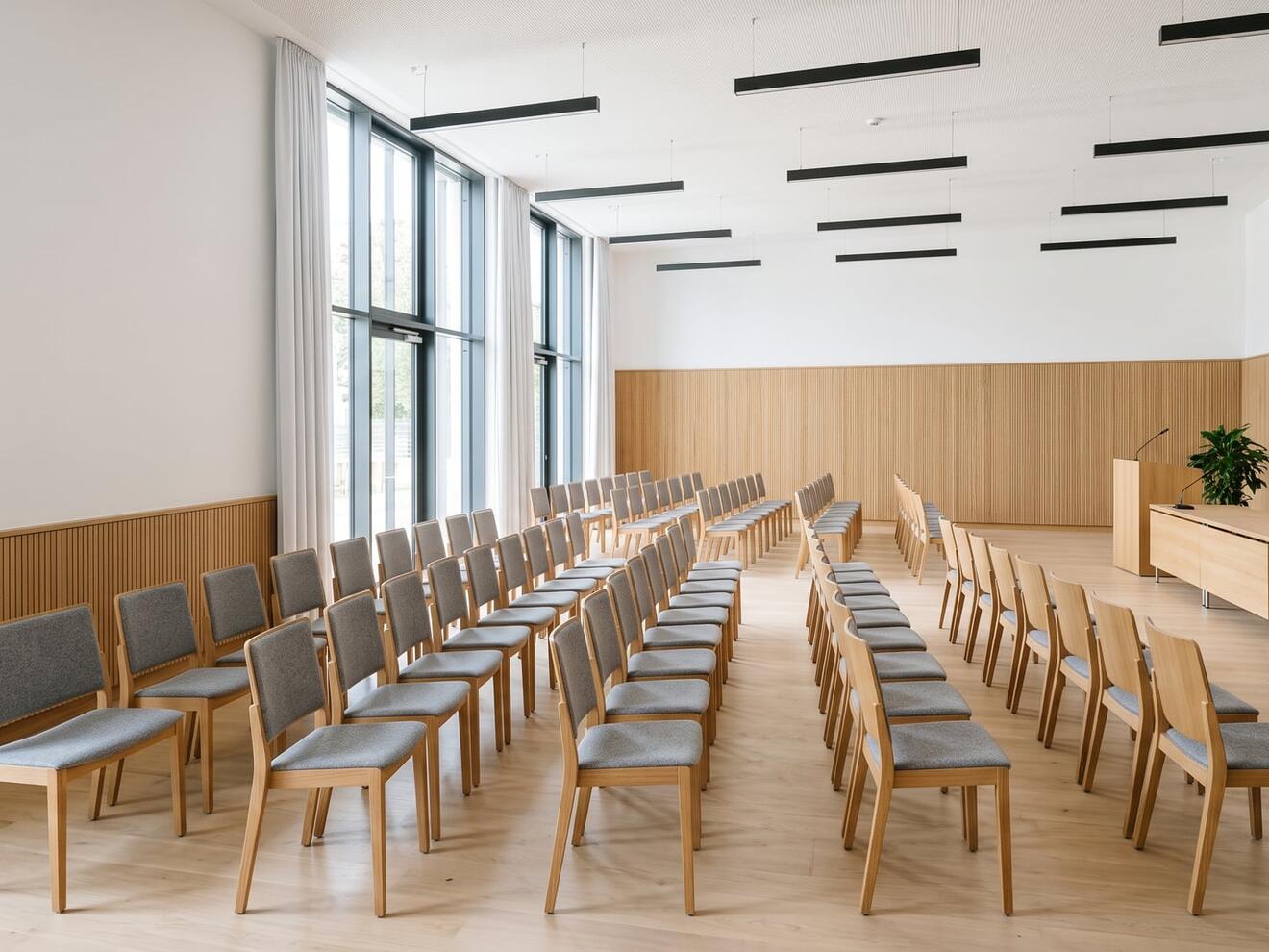 Moderner Rathaussaal mit Reihen von Stühlen, Podium und großen Fenstern.