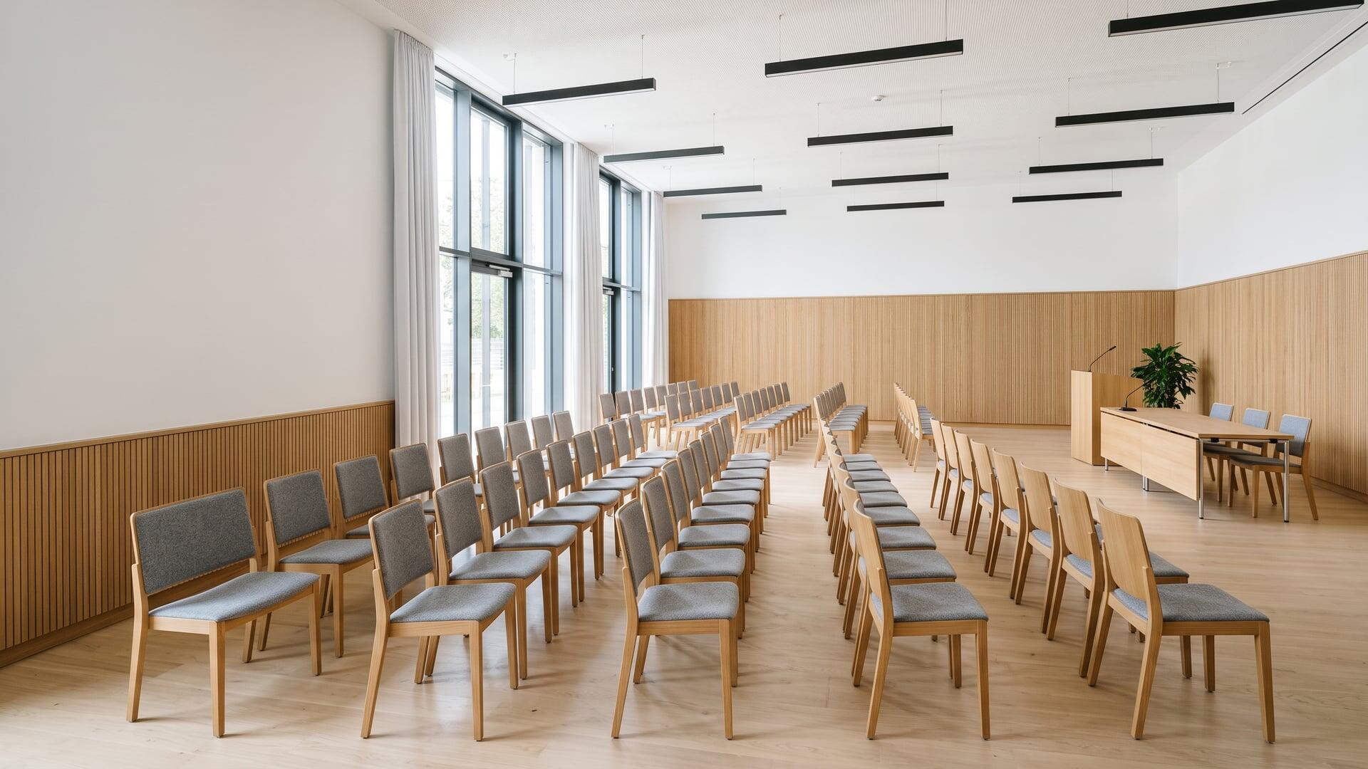 Moderner Rathaussaal mit Reihen von Stühlen, Podium und großen Fenstern.